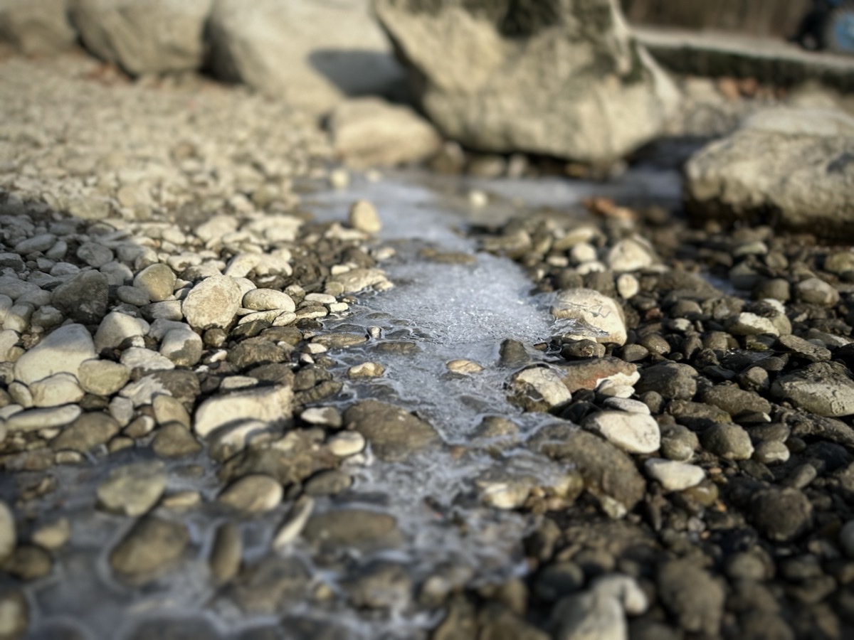 Am Flussufer liegen Kieselsteine und man sieht, wie Wasser gefroren ist.