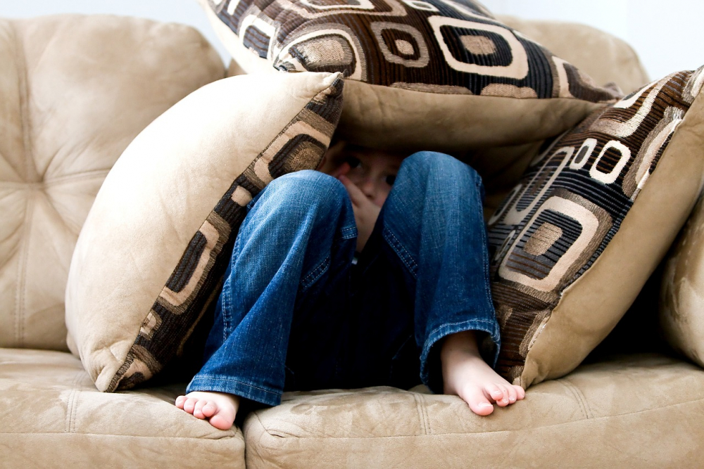 Ein Junge kauert auf einem Sofa und ist von Kissen zugedeckt. Er fürchtet sich und will sich verstecken.